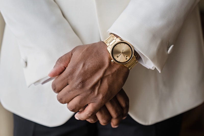 Groom wearing a crisp white suit jacket and black trousers clasps their hands behind their back, highlighting an elegant gold wristwatch which complements the formal attire at the Prestonwood Country Club.