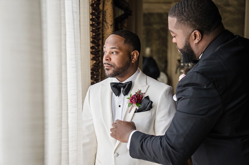 Groom in a dapper white jacket and black bowtie receives a boutonniere adjustment from another sharply dressed individual at Prestonwood Country Club, moments of sophistication captured before a grand event.