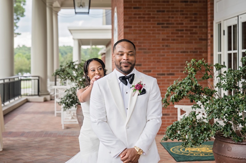 A bride playfully peeks from behind the groom, both elegantly dressed for their wedding, as they share a joyful moment on a sunny day with the charming Prestonwood Country Club as their backdrop.