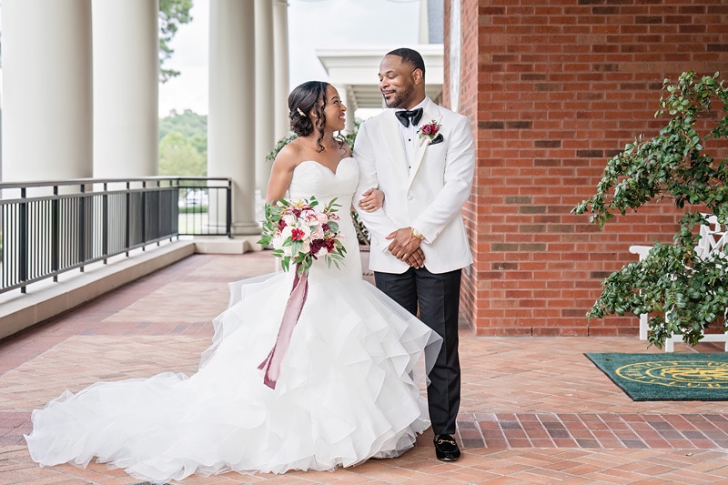 A radiant bride in a white gown with a cascading veil holds a bouquet, exchanging smiles and an affectionate gaze with a dapper groom in a black suit, amidst the classical porch setting of Preston