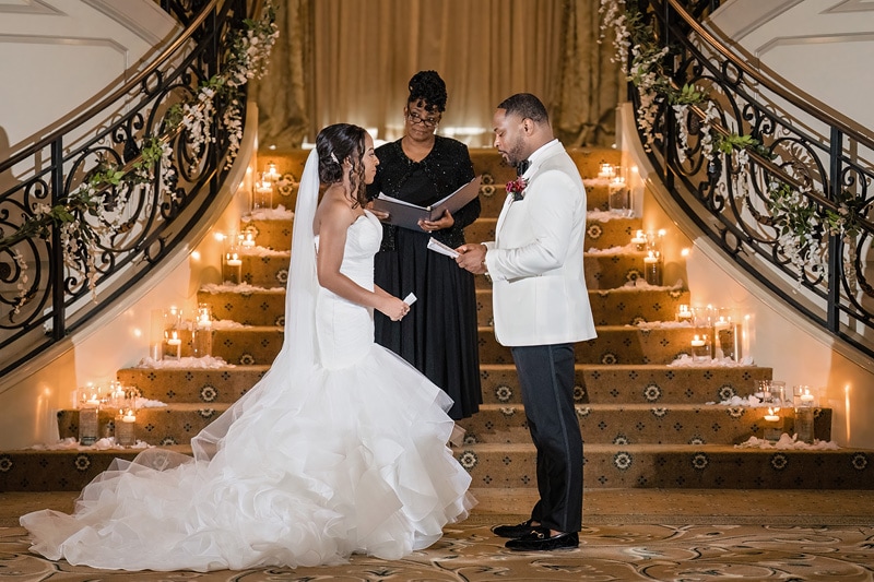 A couple exchanges vows at an elegant indoor staircase adorned with candles at Prestonwood Country Club, with an officiant leading the ceremony.