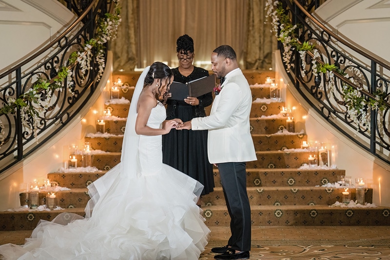 A couple exchanges vows at Prestonwood Country Club in a grand, candlelit staircase setting, officiated by a smiling celebrant, creating an intimate and elegant ambiance.