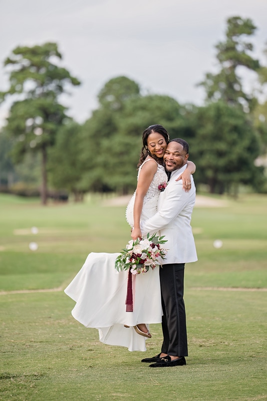 A joyful bride and groom embrace on Prestonwood Country Club's golf course, the bride being carried in the groom's arms, with a serene backdrop of lush greens and scattered trees, symbolizing their new