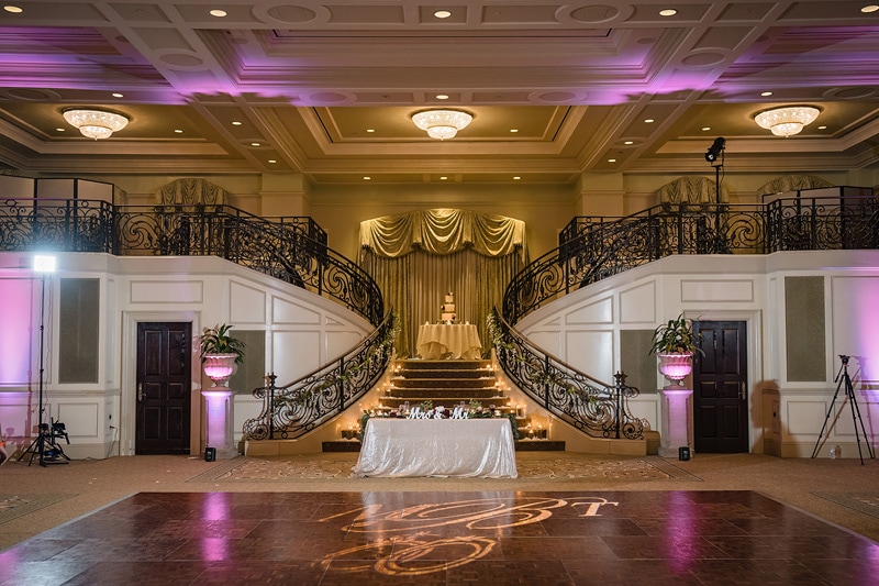 An elegant ballroom at Prestonwood Country Club, adorned with golden draperies, a grand staircase, purple ambient lighting, and a dance floor with a monogram, awaiting a festive celebration.