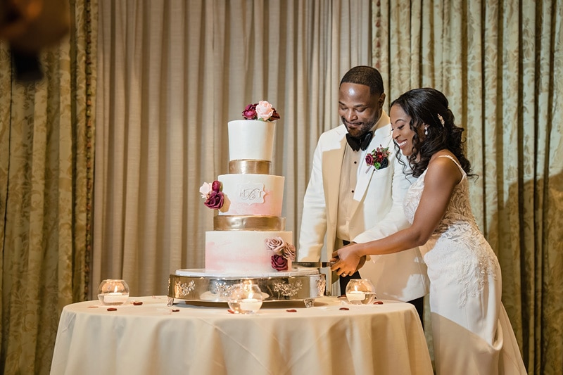A joyful bride and groom in elegant wedding attire cutting a multi-tiered cake adorned with flowers at their Prestonwood Country Club reception, surrounded by romantic candlelight.
