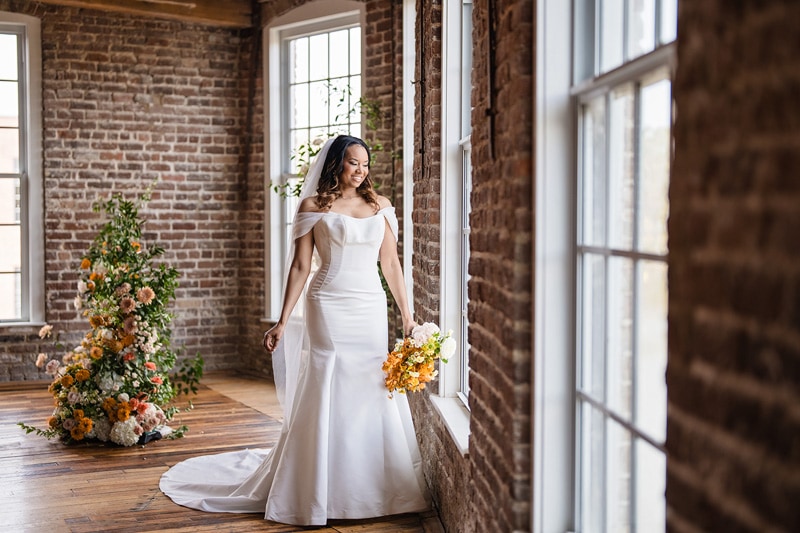 A radiant bride in an elegant off-the-shoulder gown and long veil holds a bouquet, gazing out a window in the Power House NC at Rocky Mount Mills Wedding Venue, with soft light illumin
