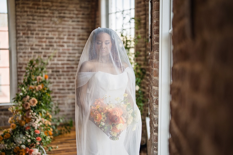 A bride in a white gown and veil holds a bouquet, standing by a window in The Power House NC at Rocky Mount Mills Wedding Venue, which is adorned with rustic brick walls and greenery.