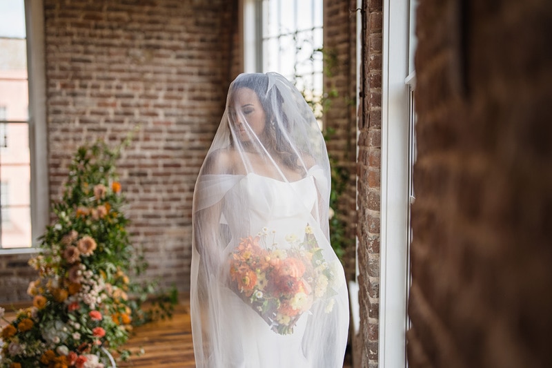 A bride in a white dress and veil holds a vibrant bouquet, pensively gazing out a window beside a brick wall at The Power House NC at Rocky Mount Mills, with soft light streaming in,