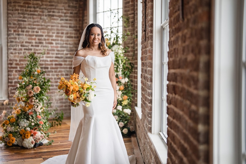 A radiant bride in an elegant off-the-shoulder white gown stands near a window in The Power House NC at Rocky Mount Mills, holding a bouquet of vibrant yellow and orange flowers, with a soft