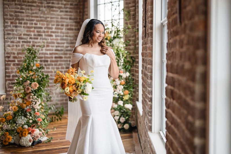 A radiant bride in a chic, off-shoulder gown holds a bouquet of yellow and orange flowers, standing beside a sunlit window at The Power House NC at Rocky Mount Mills, in a room
