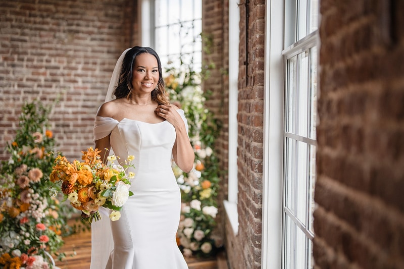 A radiant bride in an elegant off-the-shoulder gown holds a bright bouquet, her joy subtly reflected in the warm light by the window of The Power House NC at Rocky Mount Mills, a rustic