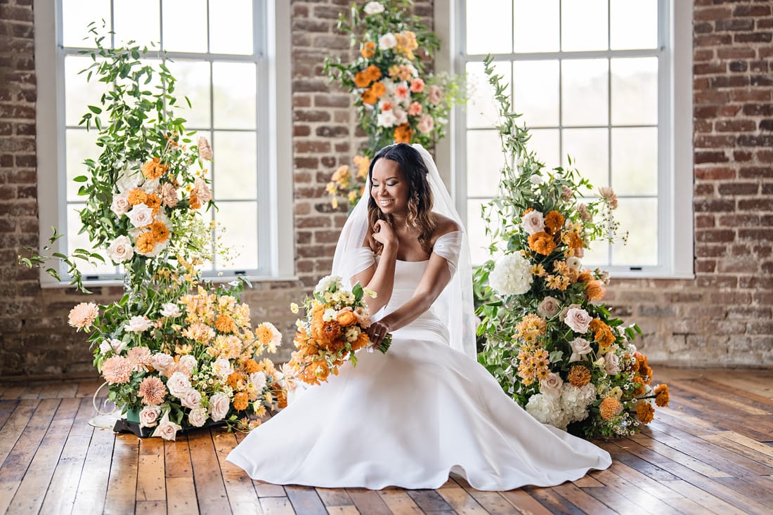 A radiant bride in a white gown sits gracefully amidst a lush arrangement of orange and white flowers at The Power House NC at Rocky Mount Mills Wedding Venue, with brick walls and large windows, basking in