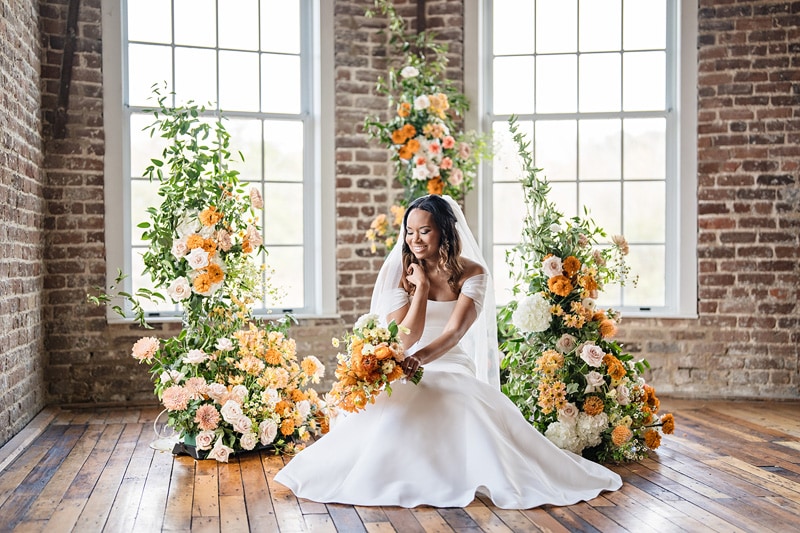 A radiant bride in a white gown sits contemplatively among cascading arrangements of peach and orange blooms in a rustic room with brick walls and large windows at The Power House NC wedding venue.