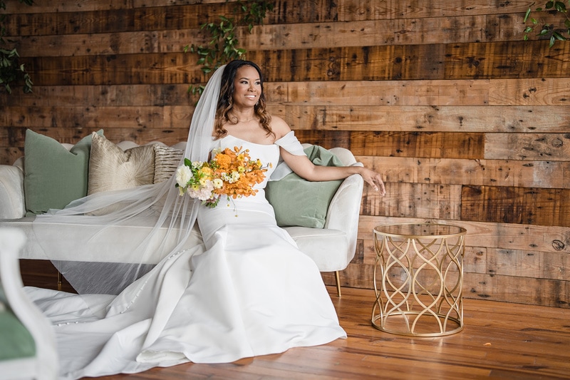 A radiant bride in a white gown and veil sits gracefully on a sofa, holding a bouquet of yellow and white flowers, at The Power House NC wedding venue, against a backdrop of warm wooden planks