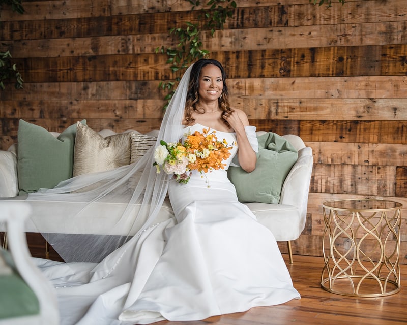 A radiant bride in a white gown sits gracefully on a couch at The Power House NC at Rocky Mount Mills, holding a bouquet of flowers, with a warm, wooden backdrop enhancing the joyous ambiance of
