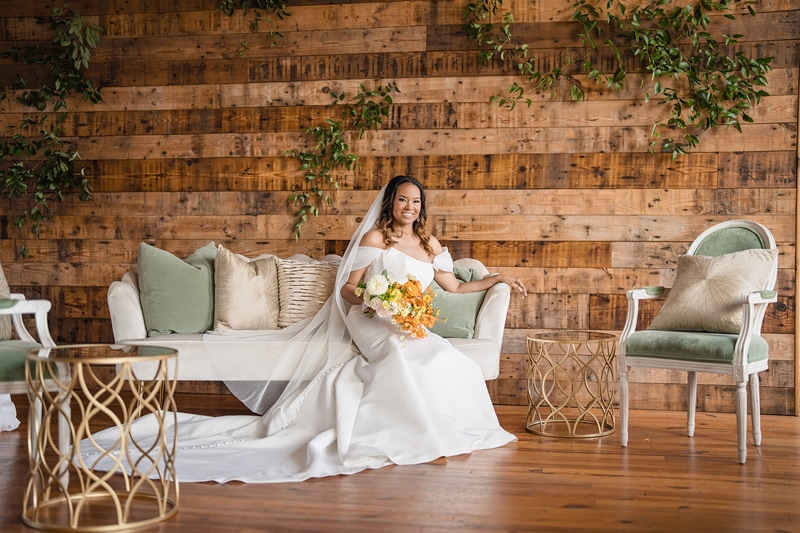 A radiant bride in a white gown, holding a bouquet, reclines gracefully on a couch in The Power House NC wedding venue, adorned with wooden walls and green vines.