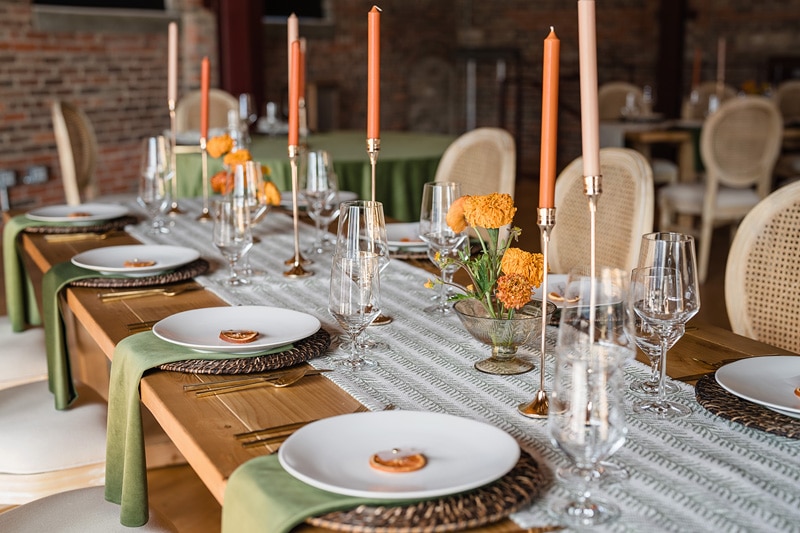 Elegant wedding dining setup with wicker placemats, white porcelain plates, and sparkling glassware, complemented by peach candles and orange floral centerpieces at The Power House NC at Rocky Mount Mills