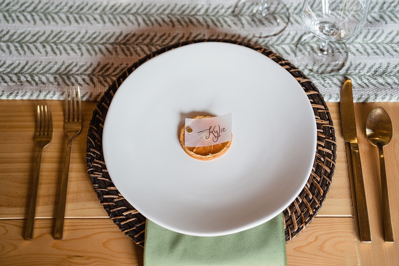 An elegant table setting at The Power House NC at Rocky Mount Mills with a white plate, golden cutlery, and a place card reading "Kyle" on a dried orange slice, resting on a