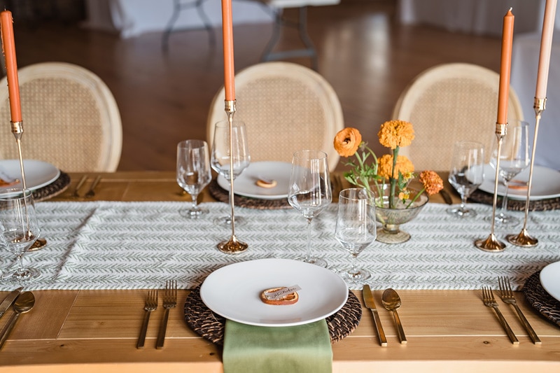 An elegantly set dining table at The Power House NC at Rocky Mount Mills, featuring a chevron runner, gold utensils, crystal glasses, woven chargers, and candles, complemented by vibrant