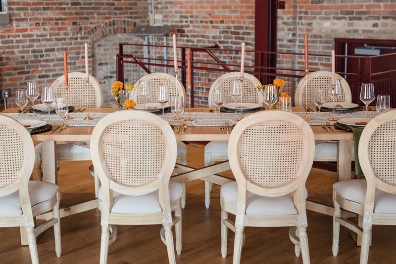 An elegantly set dining table with woven chairs, fine glassware, and vibrant yellow flowers in The Power House NC at Rocky Mount Mills, evoking a warm, inviting atmosphere for a memorable wedding venue