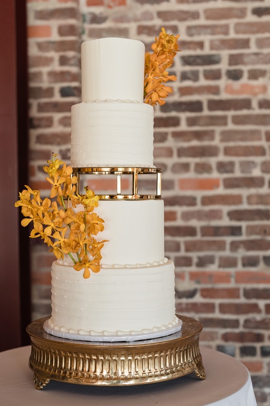 A four-tiered white wedding cake with elegant horizontal ridges, embellished with golden bands and vibrant orange flowers, presented on a golden stand against a brick wall backdrop at The Power House NC at Rocky