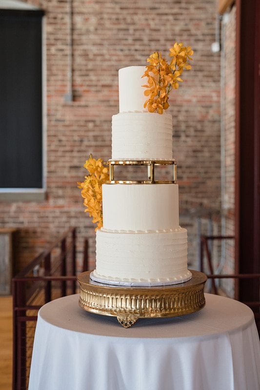 An elegant tiered wedding cake adorned with golden embellishments and yellow flowers, presented on a golden stand against a brick wall backdrop at The Power House NC wedding venue.
