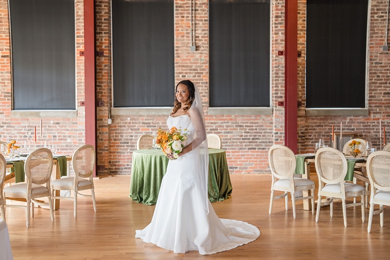 A radiant bride in a classic white gown stands poised in The Power House NC at Rocky Mount Mills, holding a vibrant bouquet, surrounded by elegantly set tables awaiting guests.