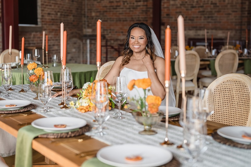 A smiling woman sits at an elegantly set table with orange flowers and candles, ready for a festive gathering in a room with rustic brick walls, perfectly capturing the essence of wedding venues in Rocky Mount NC
