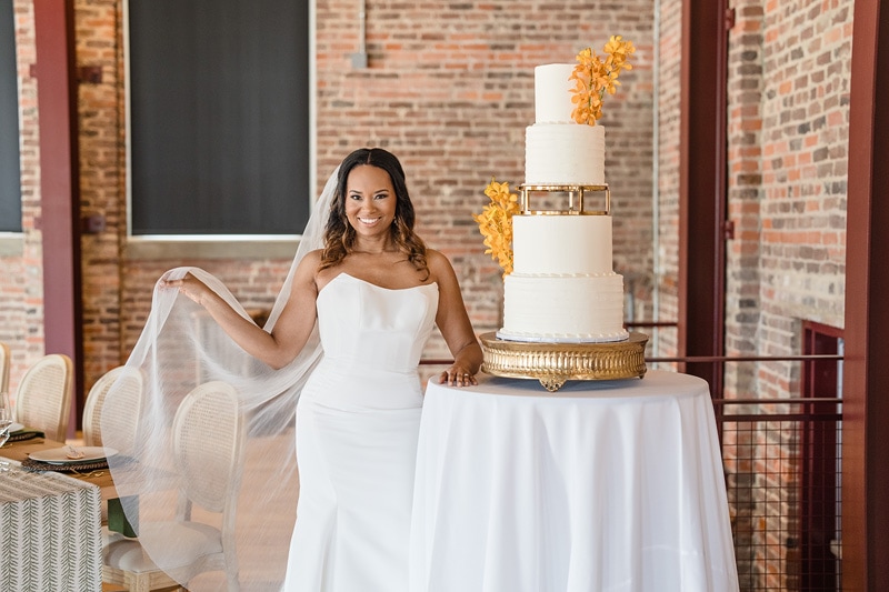 A radiant bride in a white dress with a veil stands next to a multi-tiered wedding cake adorned with golden accents and orange flowers, ready to celebrate her special day at The Power House NC wedding venue