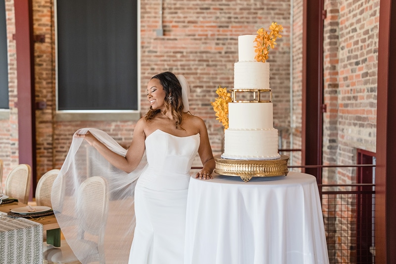A radiant bride in a white gown smiles as she unveils a grand four-tiered wedding cake decorated with gold accents and vibrant orange flowers, set against an elegant brick-walled venue at The Power House