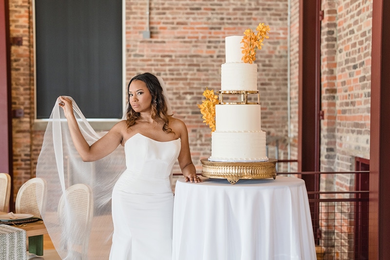 A bride in an elegant strapless gown holds her veil, contemplating a stunning four-tiered wedding cake adorned with cascading orange flowers, set in The Power House NC at Rocky Mount Mills, which ev