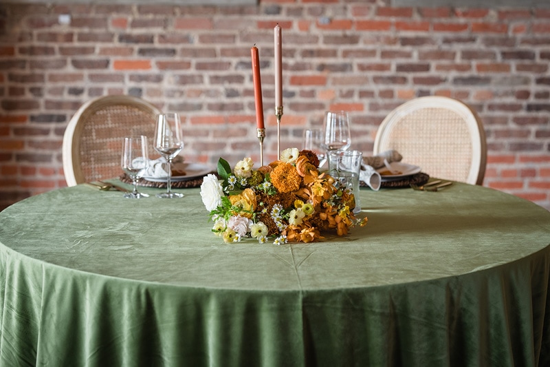 An elegantly set round dining table with a green tablecloth, floral centerpiece, tall candles, and fine dinnerware, ready for a sophisticated meal at The Power House NC wedding venue in Rocky Mount Mills