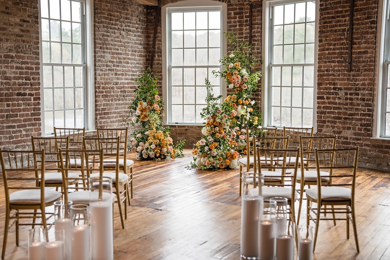 A serene wedding venue in Rocky Mount NC, with bare brick walls and hardwood floors, set with gold chairs and adorned with elegant floral arrangements beside large windows, creating a warm, inviting atmosphere.