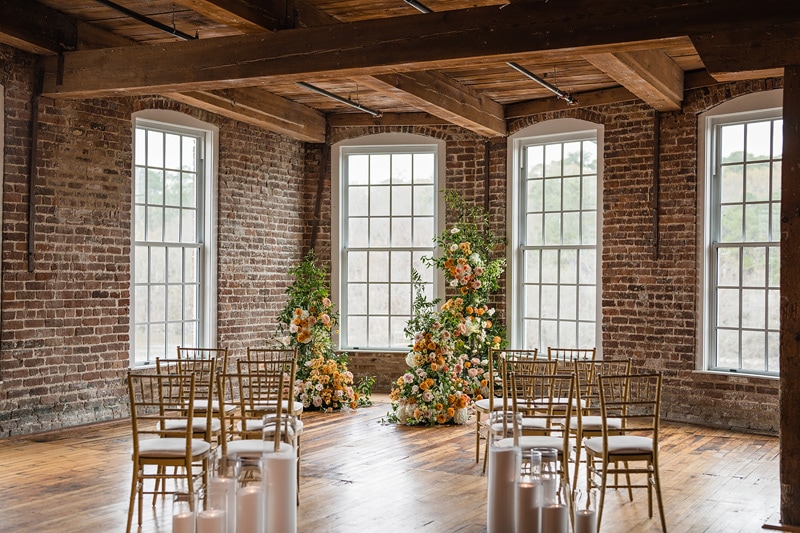 An elegant indoor wedding venue, The Power House NC at Rocky Mount Mills features exposed brick walls, wooden beams, large windows with natural light, and a floral archway beside rows of gold chiavari