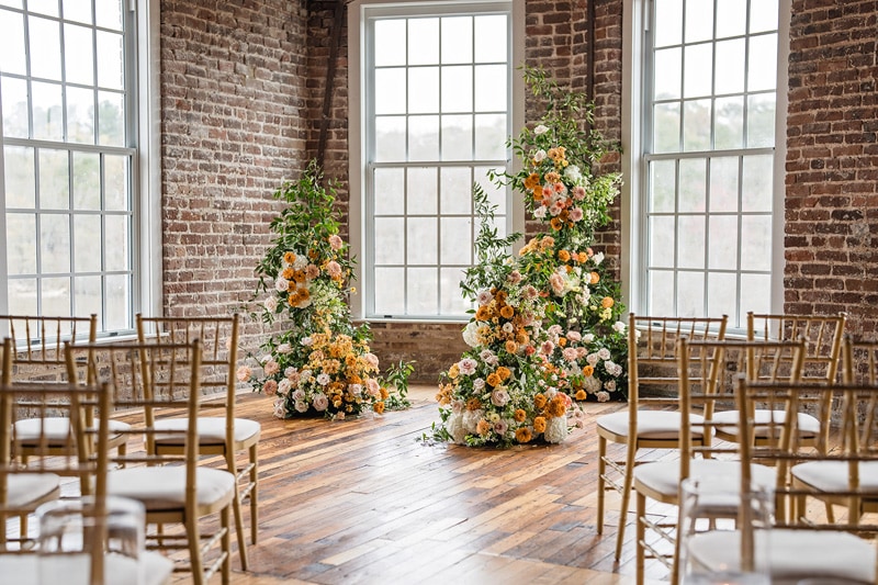 A charming wedding ceremony setup featuring elegant golden chairs and lush floral arrangements beside large windows in a rustic brick-walled venue, with sunlight filtering through, creating a warm, inviting atmosphere. This exquisite event was