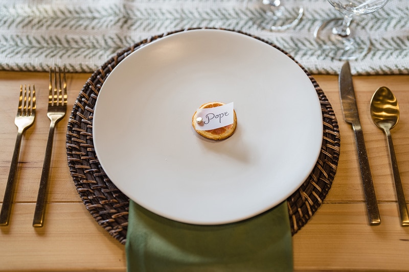 An elegantly set dining table with a wicker placemat, fine cutlery, and a pristine white plate, hosting a single dessert tagged "pope," awaiting a discerning guest at The