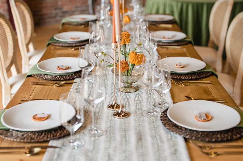 An elegantly set long dining table at The Power House NC at Rocky Mount Mills Wedding Venue, with woven placemats, white plates with napkins and rings, delicate glassware, and scattered orange