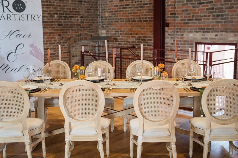 An elegant dining setup at The Power House NC at Rocky Mount Mills, arranged for a classy event, featuring fresh flowers and fine tableware, with a hair and makeup artistry sign in the background.