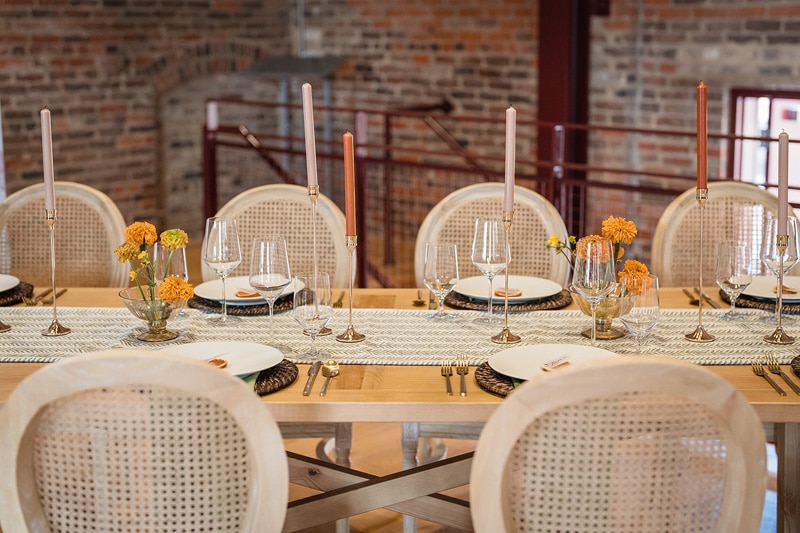 An elegantly set dining table with rattan chairs, white plates, clear stemware, and orange floral centerpieces, ready for a refined event at The Power House NC at Rocky Mount Mills, in