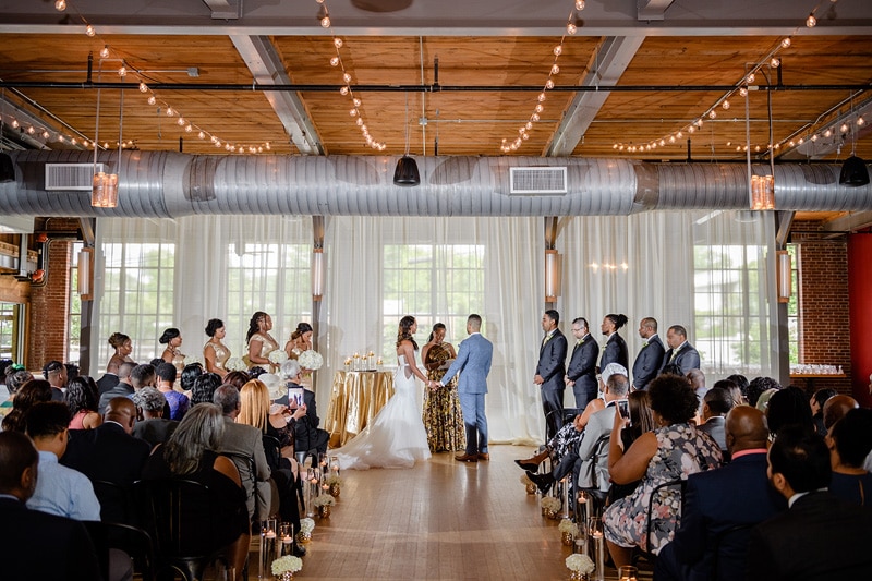 A couple exchanges vows at their wedding ceremony in an elegantly decorated industrial-chic venue, The Rickhouse, with exposed beams, string lights, and guests in formal attire witnessing the joyful moment.