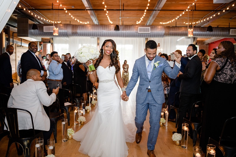 A radiant couple walks hand in hand down the aisle amidst applause at their Rickhouse wedding, the venue adorned with twinkling lights and candles, their joy is palpable.