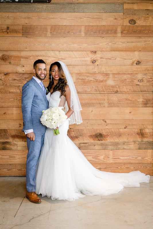 A newlywed couple poses against a wooden backdrop at The Rickhouse, the bride in a flowing white gown with a bouquet, and the groom in a tailored gray suit with a boutonniere,