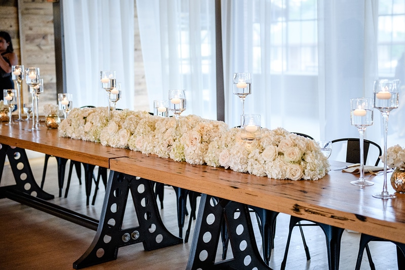 Elegant wedding table setting at The Rickhouse, featuring a lush white floral centerpiece on a rustic wooden table with whimsical polka-dotted legs, complemented by an array of glowing candles, set