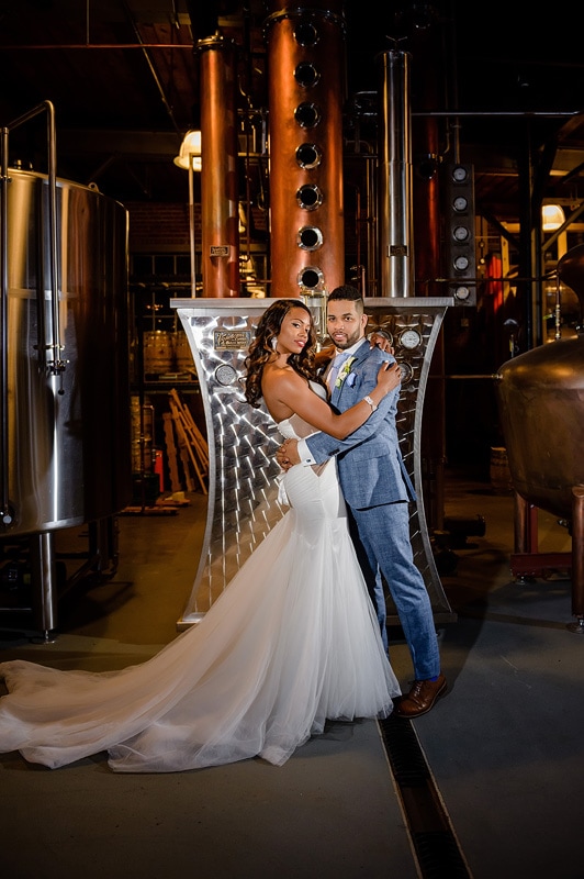 A happy couple in wedding attire sharing an intimate moment amidst the industrial backdrop of The Rickhouse, with stainless steel tanks and copper pipes enhancing the unique charm of their special day.
