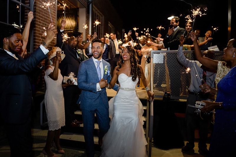 A joyful bride and groom exit through a sparkling archway of sparklers held by friends and family at The Rickhouse, celebrating the couple's wedding night with wide smiles and festivity.