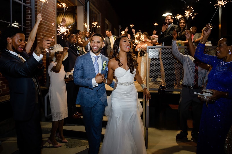 A joyful newlywed couple walks hand-in-hand through a sparkling tunnel of friends and family holding sparklers, celebrating their marriage at The Rickhouse wedding reception, an evening outdoor event.