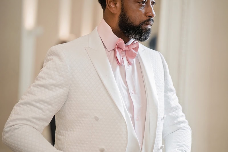 A distinguished man with a beard wearing a white textured suit buttoned properly according to the suit button rule, with a pastel pink shirt and bow tie, looking thoughtfully to his left in a room