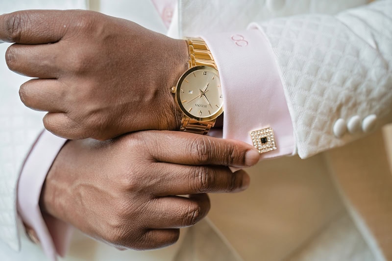 Close-up of a person's hands adjusting their cufflink, wearing a pink shirt and a three-button white patterned suit. One hand displays a gold wristwatch. Elegance and attention to detail