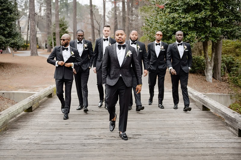 A group of six men in elegant black two-button tuxedos with boutonnieres, confidently walking along a wooden bridge in a park, with trees lightly blurred in the background. The man in
