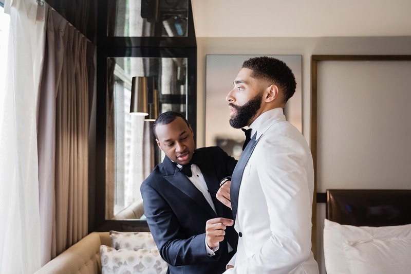 A groom in a white shirt looks into a mirror, as another man in a black three-button suit adjusts the groom's bow tie in a stylishly furnished room with modern decor.